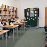 drużyny siedzące przy stolikach i uczniowie oglądający konkurs w czytelni szkolnej