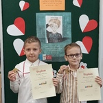 Uczeniowie z dyplomami i medalami za zajęcie 1 miejsca w konkursie na tle tablicy z tytułem konkursu i plakatem i serduszkami w biało-czerwonych barwach