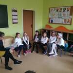 Pani czyta książkę, dzieci siedzą w półkolu na krzesełkach