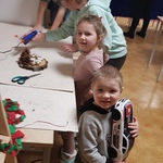 na pierwszym planie dwoje młodszych dzieci stoi przy stoliku, chłopiec trzyma zabawkę samochód, w oddali starsze dzieci i rodzice