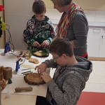 przy stoliku siedzi jeden uczeń i coś przykleja do okrągłej podstawki z pieńka, za nim stoi kobieta, dalej kolega pochylony nad stroikiem, który wykonuje