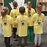 uczniowie w koszulkach z logo szkoły, odwróceni do regału prezentują tył koszulki