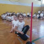 Dzieci na sali gimnastycznej siedzą w rzędach przygotowane do zawodów sportowych